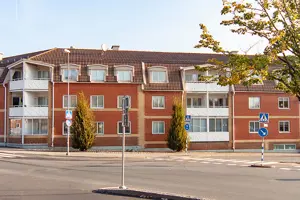 Flerbostadshus med röd tegelfasad, vita fönster och rött tegeltak, träd i förgrunden | Nivika Fastigheter AB (publ)