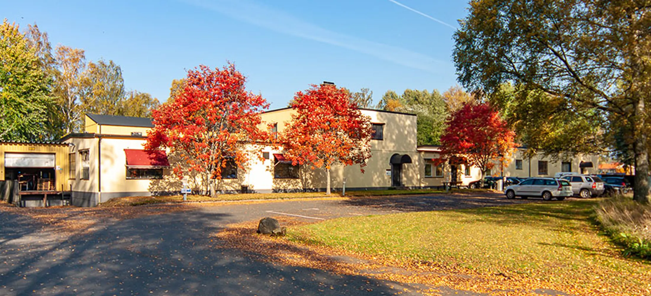 Kommersiell fastighet med gul fasad, röda markiser och tre träd på framsidan | Nivika Fastigheter AB (publ)